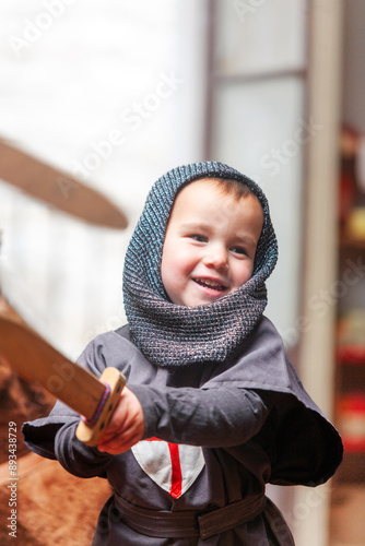 A kid is dressed in a knight costume and holds a wooden sword while playing smiling in the playroom of his home.