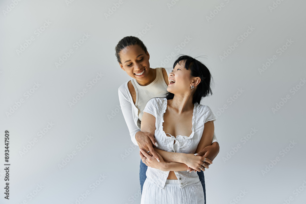 Two young women, one Black and one Asian, laugh as they hug each other in front of a grey background. Two young women, one Black and one Asian, laugh as they hug each other in front of a grey background.