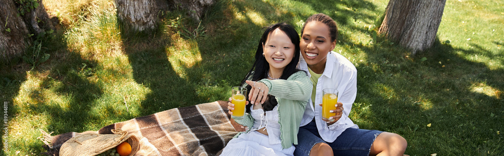 Two young women, one African American and one Asian, share a picnic on a grassy park blanket, smiling and enjoying each others company Two young women, one African American and one Asian, share a picnic on a grassy park blanket, smiling and enjoying each others company