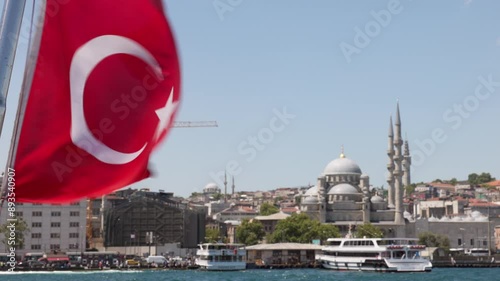 A Turkish flag waves in the wind with Istanbul's skyline in the background, including a mosque and boats on the water.
