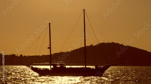 A sailboat sails across the water at sunset, with a hillside silhouetted in the background. Sailing boat at sunset.
