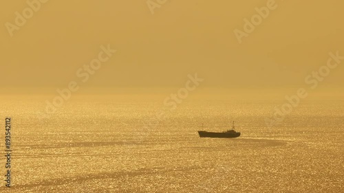 A lone ship sails across the glistening sea as the sun sets, casting a golden glow on the water. Ship sailing on golden sea at sunset