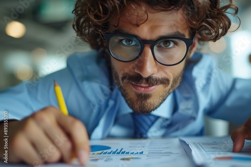 A man with curly hair and glasses is focusedly analyzing charts and graphs, holding a pencil, suggesting a professional and analytical environment. photo