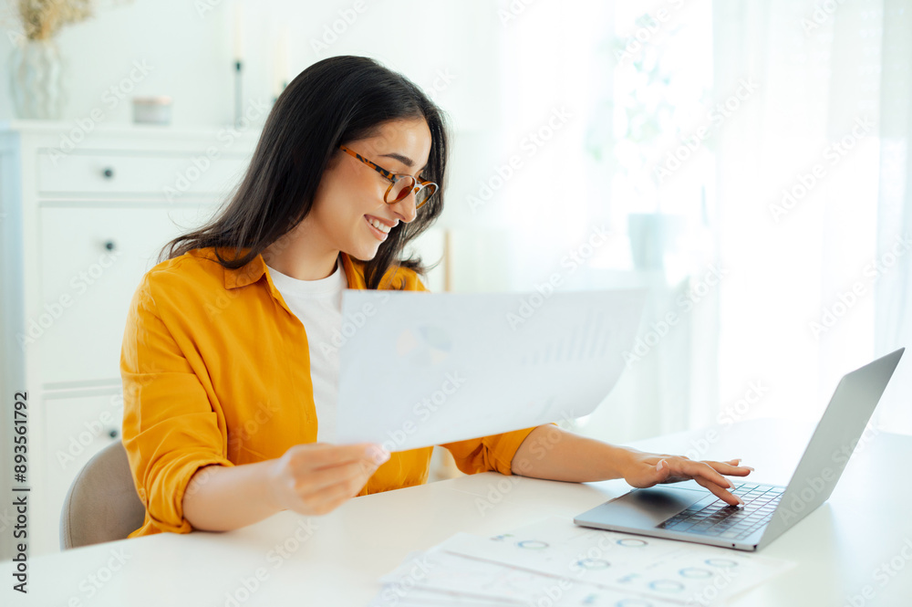 Attractive concentrated arabian or indian brunette woman, wearing eyeglasses, secretary or financial manager, sits at a table with laptop, holding documents in hands, searching information at a laptop