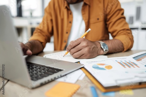 A man is focused on analyzing business strategy at his desk, using a laptop, charts, and paperwork. He is professionally working on data analysis with concentration in a modern office setting