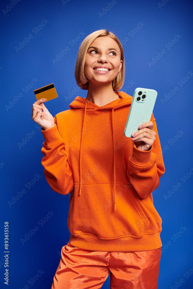 beautiful joyous woman with blonde hair in vibrant orange hoodie posing with credit card and phone beautiful joyous woman with blonde hair in vibrant orange hoodie posing with credit card and phone