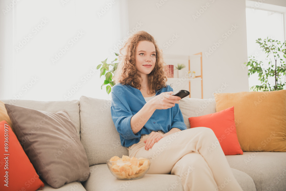 Photo of attractive trendy young woman sit sofa watch tv eat chips blue shirt move new apartment home interior indoors Photo of attractive trendy young woman sit sofa watch tv eat chips blue shirt move new apartment home interior indoors