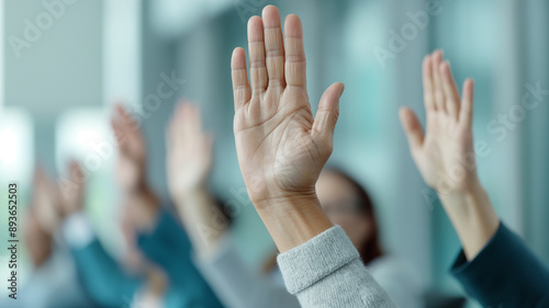 Close-up of multiple hands raised during an office meeting, indicating active participation and engagement. The background shows a professional setting with blurred attendees.