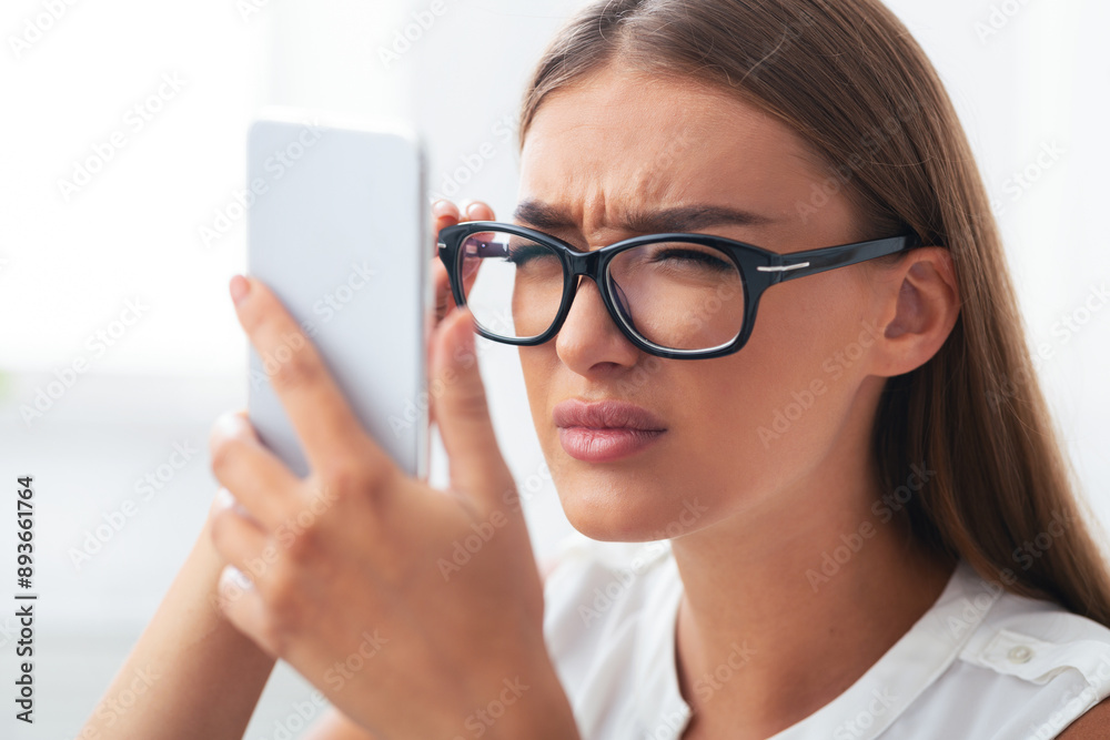 Poor Eyesight. Girl Squinting Eyes Looking At Smartphone Screen Through Eyeglasses Sitting Indoor. Selective Focus Poor Eyesight. Girl Squinting Eyes Looking At Smartphone Screen Through Eyeglasses Sitting Indoor. Selective Focus