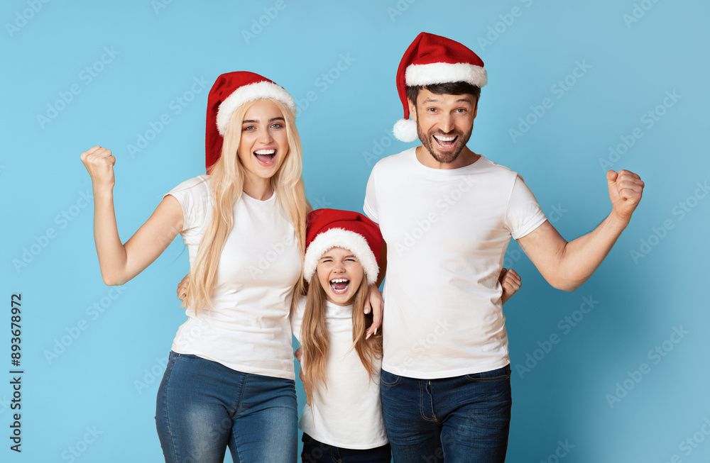 Obraz premium A family of three, wearing Santa hats and white t-shirts, pose with their arms raised in the air and smiling excitedly in front of a blue background.