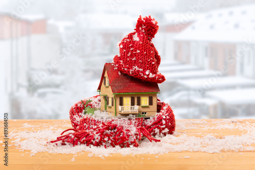 thermal insulation: old model house wrapped up warm with red scarf and bobble hat, cosy lit windows against a backdrop of snow-covered terraced houses in winter