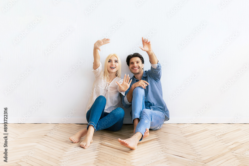 A young couple sits on a hardwood floor with their arms raised. A young couple sits on a hardwood floor with their arms raised.