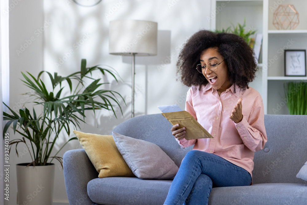 Happy woman receiving exciting news from letter while sitting comfortably on living room sofa. Brightly lit interior with modern design and plants adds warmth and positivity to the scene. Happy woman receiving exciting news from letter while sitting comfortably on living room sofa. Brightly lit interior with modern design and plants adds warmth and positivity to the scene.