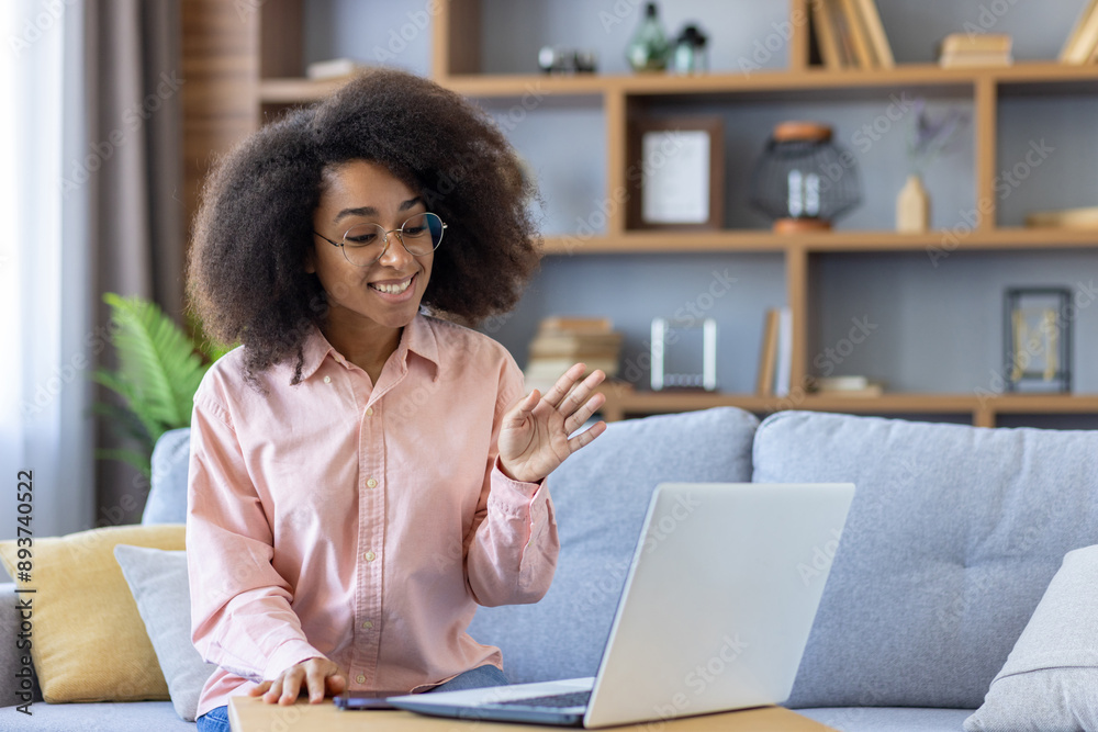 Smiling woman with curly hair participates in a video call, waving at the screen while sitting on a comfortable couch with laptop in modern living room Smiling woman with curly hair participates in a video call, waving at the screen while sitting on a comfortable couch with laptop in modern living room