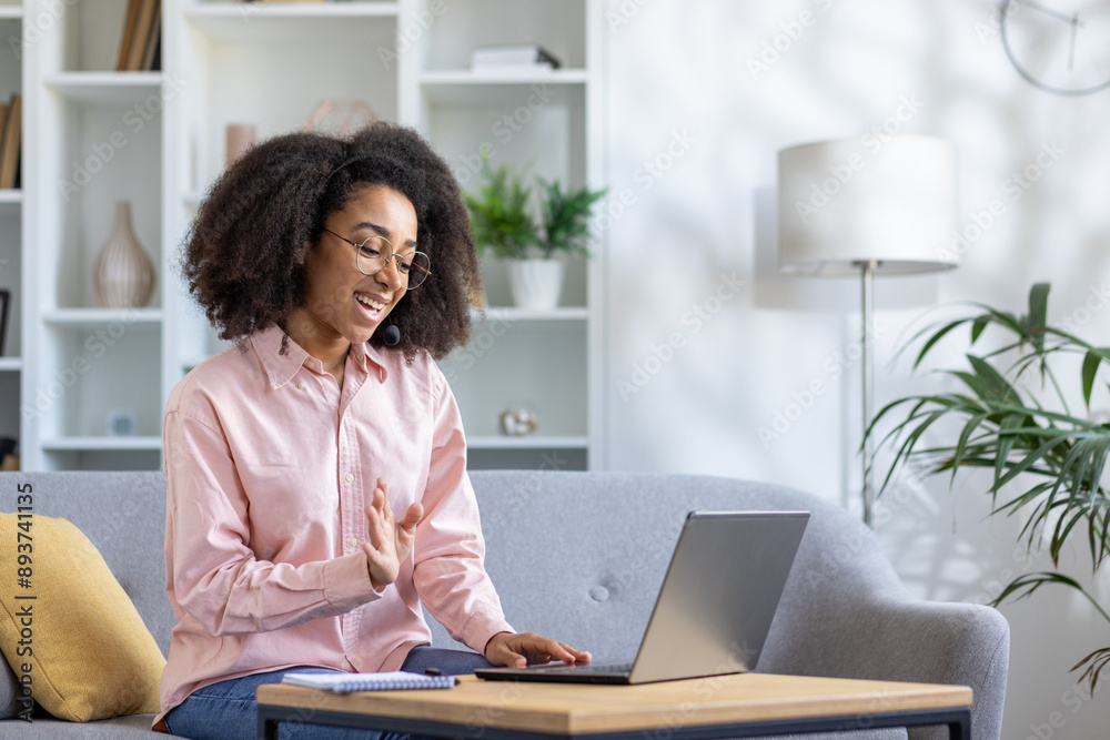 Smiling young woman working from home on laptop wearing headphones in cozy modern living room. Woman gesturing during online meeting, engaging in virtual communication. Smiling young woman working from home on laptop wearing headphones in cozy modern living room. Woman gesturing during online meeting, engaging in virtual communication.