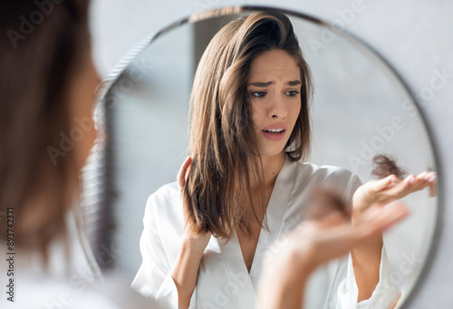 Hairloss Concept. Worried Young Woman Holding Bunch Of Fallen Hair In Hand While Standing Near Mirror In Bathroom, Stressed Beautiful Lady Suffering Alopecia Or Health Problems, Selective Focus photo