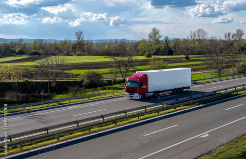 A big red truck with a white trailer drives on the highway through the rural areas of Serbia. Beautiful blue sky with white clouds above the road.
