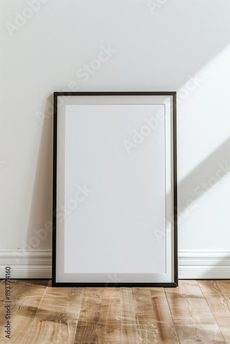 A blank white picture frame with a black border resting against a white wall on a wooden floor.