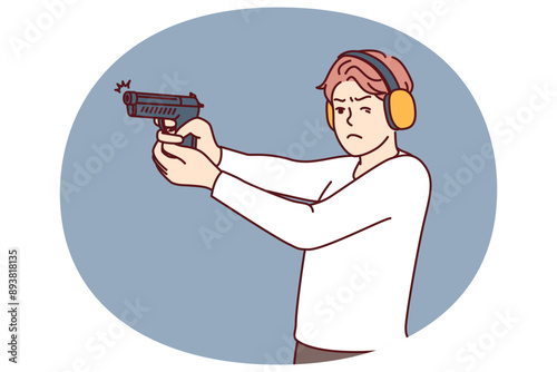 Man with gun trains at shooting range to learn how to shoot and become sheriff or bodyguard. Guy in ballistic headphones holds gun and squints to hit target during training in shooting range