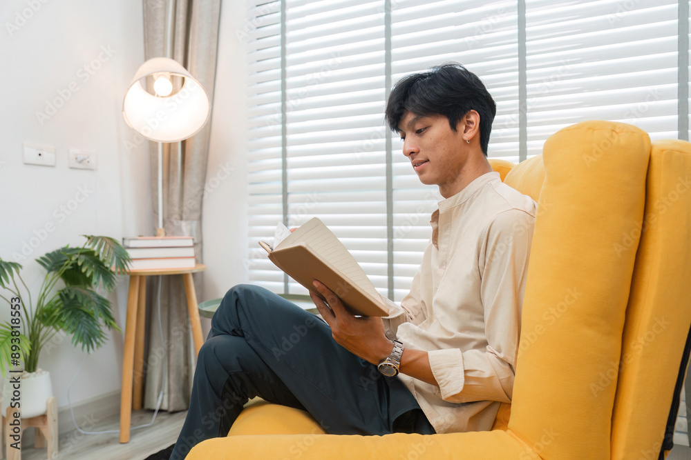 Young Man Relaxing in Modern Living Room, Reading a Book on a Comfortable Yellow Chair Young Man Relaxing in Modern Living Room, Reading a Book on a Comfortable Yellow Chair