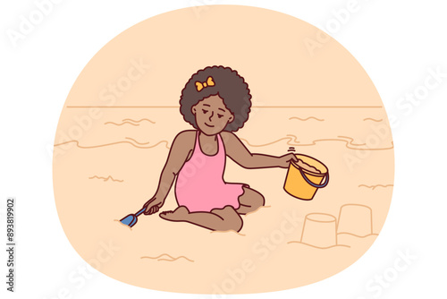 Little girl playing with sand sitting on beach building castle with bucket and shovel during summer holidays at resort. Pre-teenage girl located on sea beach relaxing alone on sea or ocean