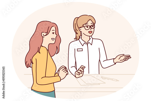 Woman working at reception in hotel or beauty salon consults client, helps to choose beautician and best procedure. Girl visiting cosmetology clinic stands near reception with administrator
