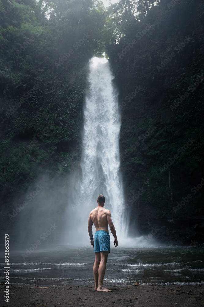 Fototapeta premium Homme qui se tient près d'une grande cascade d'eau, en Indonésie, à Bali, dans une nature luxuriante.