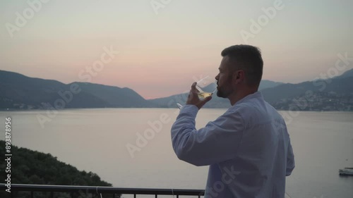 Kotor Bay, Montenegro. Handsome man drinking white wine standing on the patio of the luxury hotel and looking at the sunset, sea and mountain