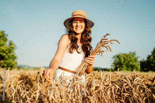 One beautiful young caucasian woman wearing white dress and straw hat holding crops of wheat and standing in wheat field during the day 