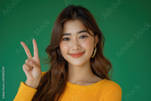 Happy young Asian woman smiling and showing victory sign wearing yellow shirt on green background, 