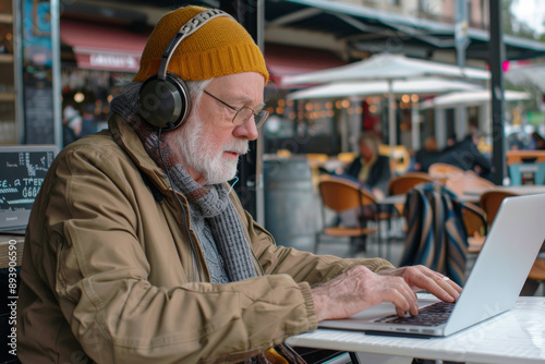 Another elderly man, bundled up in a beanie and scarf, works on his laptop with noise-cancelling headphones in a bustling outdoor café.