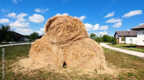 A stack of three round hay bales sits in a village yard, with a path leading to the houses in the background. Beautiful rustic scenery