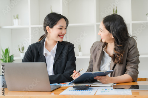 Collaborative Synergy: Two young Asian businesswomen engage in a productive meeting, their smiles reflecting shared success and teamwork in a bright office setting. 