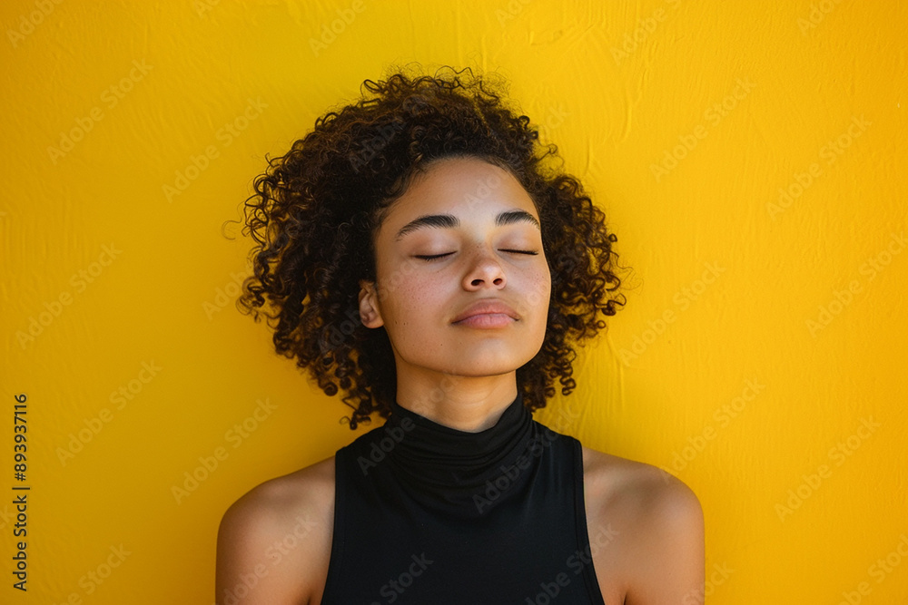A young woman with curly hair standing against a bright yellow background with her eyes closed, representing relaxation and mindfulness. The vibrant color adds a lively contrast.