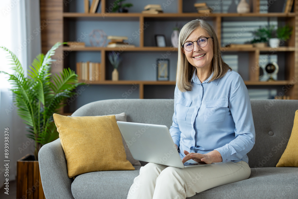 Smiling senior woman working on laptop sitting on sofa. Comfortable home setting with bookshelves and plants in background. Concept of remote work, technology, senior lifestyle, happiness. Smiling senior woman working on laptop sitting on sofa. Comfortable home setting with bookshelves and plants in background. Concept of remote work, technology, senior lifestyle, happiness.