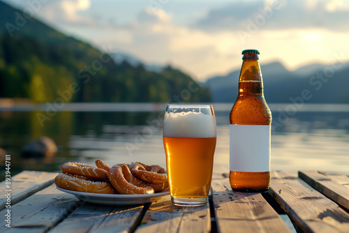 A close up of a bottle of beer with a blank mock up label on it and a glass of beer served together with brezels on the wooden table with a lake at sunset in the background photo