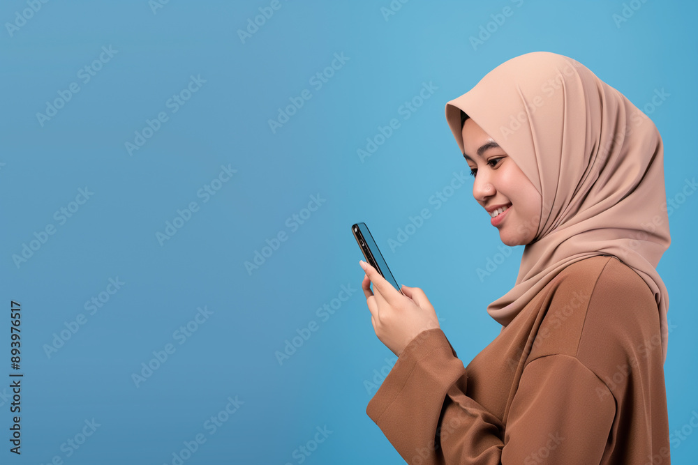 Smiling young woman in beige hijab using smartphone, isolated against blue background, modern communication and technology concept, joyful and connected