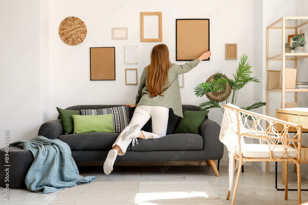 Young woman hanging picture on white wall near sofa in living room Young woman hanging picture on white wall near sofa in living room