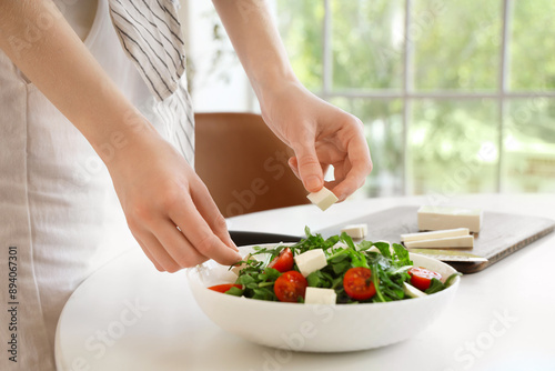 Young woman putting pieces of feta cheese in salad at home