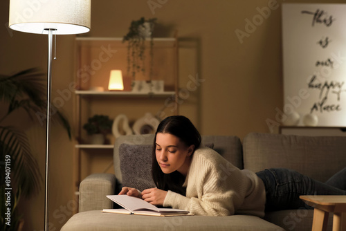 Young woman reading book and resting on cozy sofa in living room at night photo