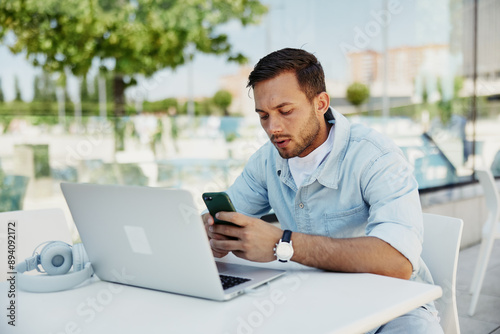 Young man working outdoors on laptop, checking phone notifications, focused on multitasking