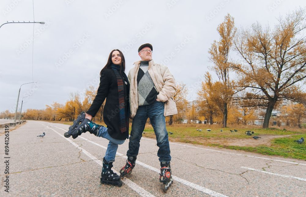 An elderly father in a down jacket, jeans and roller skates stand near his young funny daughter in a coat and roller skates. The concept of understanding between parents, children and happy retirement An elderly father in a down jacket, jeans and roller skates stand near his young funny daughter in a coat and roller skates. The concept of understanding between parents, children and happy retirement