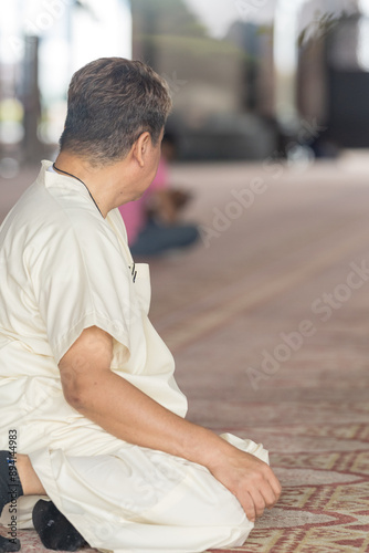 Fifty-something Muslim Malay person in religious attire, wearing a white outfit and a black hat, praying devoutly on a majestic carpet inside a very large mosque in Putrajaya, Malaysia.