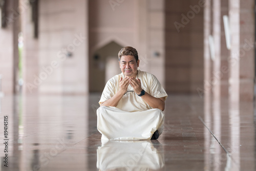 Fifty-something Muslim Malay person in religious attire, wearing a white outfit and black hat, praying devoutly in a wide glass-enclosed marble area outside very large mosque in Putrajaya, Malaysia. photo