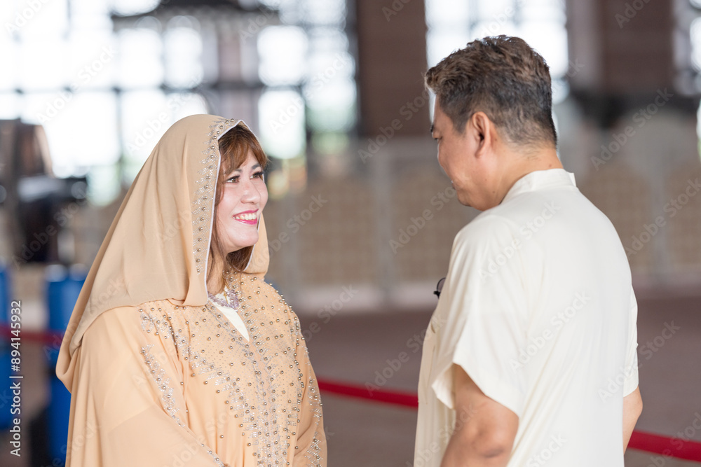 50-something Muslim Malay man in white outfit and black hat and 30-something Chinese woman in cream-colored religious attire standing together inside large mosque in Putrajaya, Malaysia. 50-something Muslim Malay man in white outfit and black hat and 30-something Chinese woman in cream-colored religious attire standing together inside large mosque in Putrajaya, Malaysia.