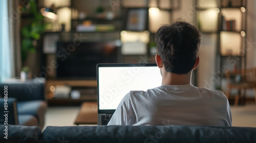 Young man seen from behind using a laptop with an empty white screen, in a home setting