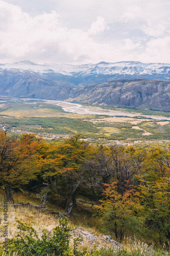 A mountain range with a river running through it. The trees are orange and yellow, and the sky is cloudy A mountain range with a river running through it. The trees are orange and yellow, and the sky is cloudy
