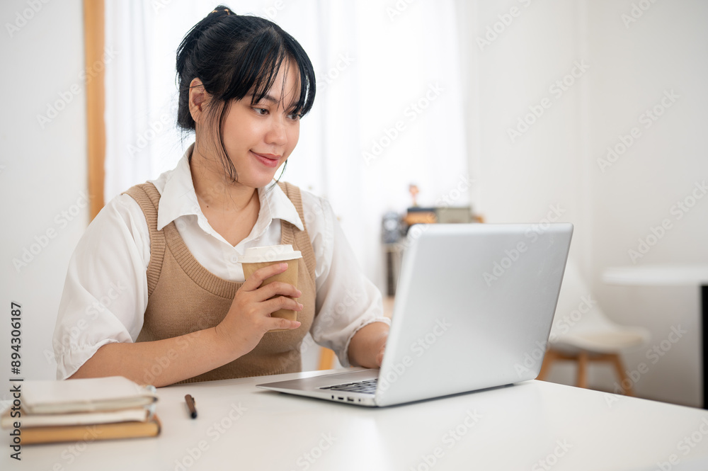 An Asian woman working remotely from a cafe, working on her laptop computer and drinking coffee. An Asian woman working remotely from a cafe, working on her laptop computer and drinking coffee.