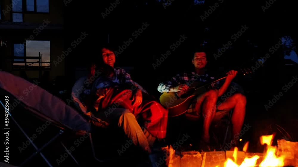 The family is sitting around the campfire, man plays the guitar, a woman listens and sings along, my daughter wrapped herself in a blanket. Family summer evening in the courtyard of the house The family is sitting around the campfire, man plays the guitar, a woman listens and sings along, my daughter wrapped herself in a blanket. Family summer evening in the courtyard of the house