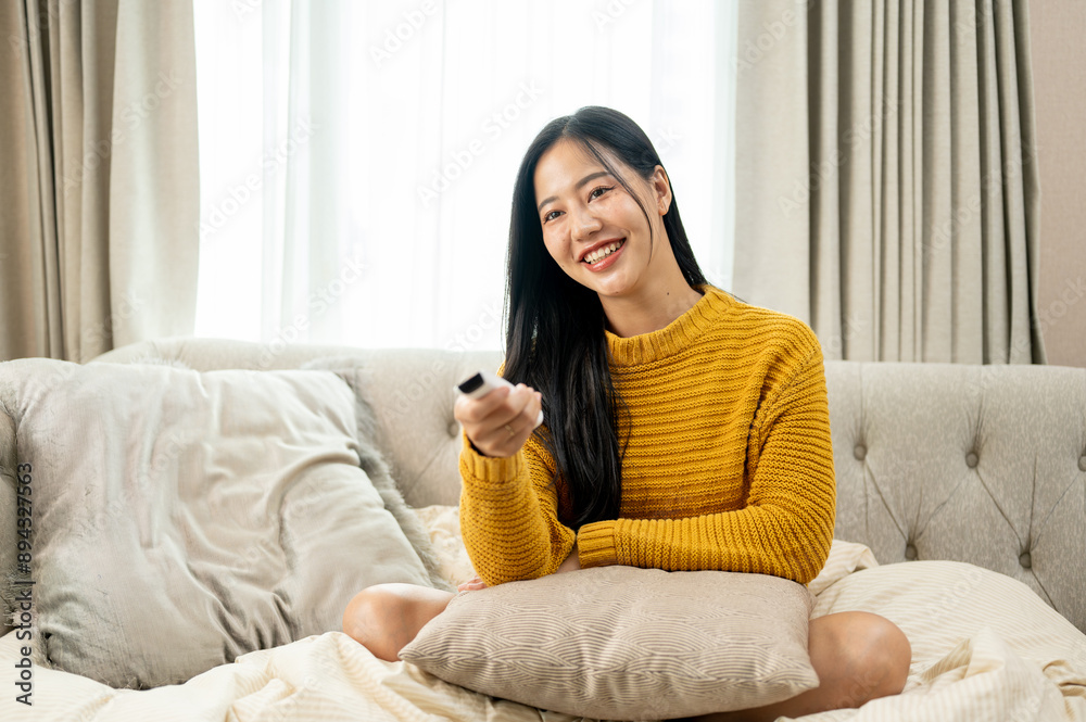An attractive Asian woman is watching TV on a couch in the living room, holding a TV remote control. An attractive Asian woman is watching TV on a couch in the living room, holding a TV remote control.